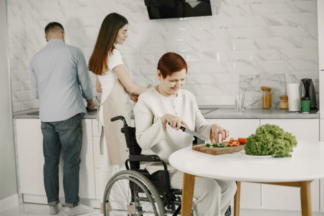 Une femme en fauteuil roulant coupe des légumes dans une cuisine accompagnée de deux autres personnes
